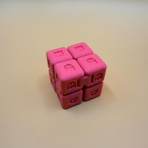 May include: A pink cube-shaped fidget toy featuring a ghost design on each face. The toy is made of a soft material with a smooth texture. The cube is composed of smaller cubes that can be rotated and manipulated.