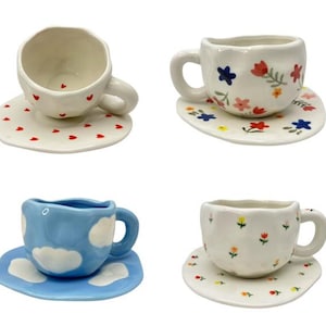 May include: Four ceramic teacups and saucers. One cup is white with red hearts, another is white with colorful flowers, a third is blue with white clouds, and the last is white with small flowers. Each saucer matches the cup's design.