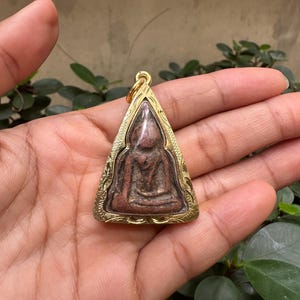 Puede incluir: Un colgante triangular con una figura de Buda sentado. El colgante tiene un marco dorado con detalles intrincados en los bordes. La figura de Buda es de color marrón rojizo.