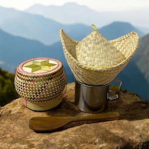 Könnte beinhalten: Ein geflochtener Bambus-Reiskocher mit Deckel und einem Holzlöffel. Der Kocher steht auf einem Stein mit einer Metalltasse daneben.