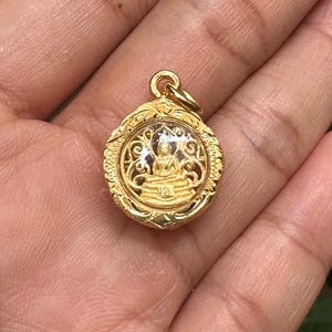 Könnte beinhalten: Goldfarbener Anhänger mit einer sitzenden Buddha-Figur in einem transparenten, kreisförmigen Rahmen. Der Anhänger hat einen verzierten, strukturierten Rand und eine kleine Schlaufe für eine Kette. Der Buddha befindet sich in meditativer Pose.