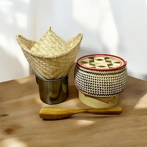 Può includere: Un set di tre cesti in bambù intrecciato, tra cui un cesto rotondo con un coperchio a motivi rosso e bianco, un cesto conico alto con un bordo largo e una paletta di legno.