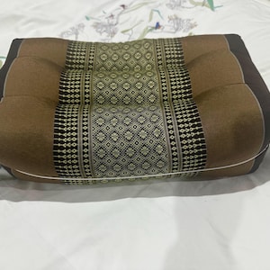 May include: A square, brown cushion with a central patterned panel in olive green and black. The cushion has a tufted design and is placed on a white surface. The cushion's edges are a darker brown.