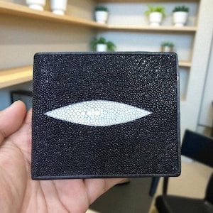 Könnte beinhalten: Schwarzes Lederportemonnaie mit einem markanten weißen Stechrochen-Augen-Design. Das Portemonnaie hat eine strukturierte Oberfläche und ein zweifach gefaltetes Design. Das augenförmige Detail ist mittig auf der Vorderseite.