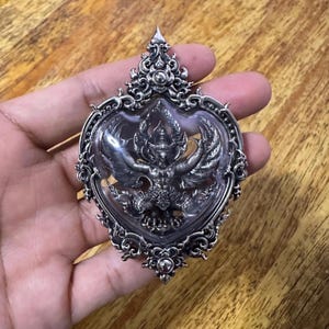 May include: Ornate silver pendant featuring a detailed Garuda figure within a heart-shaped frame. The pendant has intricate filigree and a clear, protective covering over the central design. The pendant is held in a hand against a wooden background.