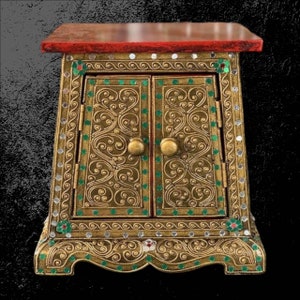 May include: An ornate, gold-coloured cabinet with two doors, featuring intricate scrollwork and green and white accents. The top is a deep red, and the cabinet has small, round gold knobs. A decorative piece.