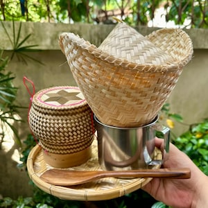 May include: A set of woven bamboo kitchenware, including a rice steamer, a small bowl, a wooden spoon, and a metal cup. The items are arranged on a woven bamboo tray.