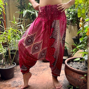 Puede incluir: Pantalones tipo harén holgados en un tono magenta intenso con un patrón repetitivo de ojos de plumas de pavo real en negro, blanco y dorado. Los pantalones tienen una cintura alta elástica fruncida con cordón y puños elásticos en los tobillos.