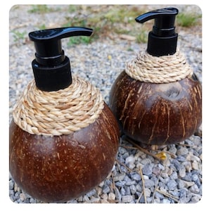 Puede incluir: Dos dispensadores de jabón de cáscara de coco marrón con bombas negras y detalles de cuerda de fibra natural. Los dispensadores están sentados sobre un lecho de pequeñas piedras grises.