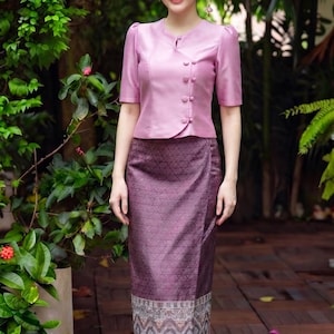 May include: A two-piece outfit consisting of a light pink short-sleeved blouse with a round neckline and a side button closure, paired with a long, dark purple wrap skirt featuring a geometric pattern and a detailed woven border at the hem.