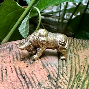 May include: A small, gold-colored metal figurine of a bull standing on a textured, brown surface. The bull is facing left and has its head lowered as if it is grazing.