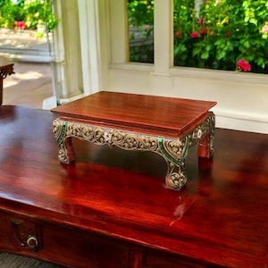 May include: A small, ornate wooden table with a dark red finish. The table has intricate carvings and gold accents. The table is sitting on a larger, dark red table.