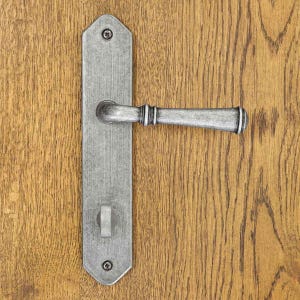 Peut inclure: Une poignée de porte en métal argenté avec une plaque rectangulaire aux extrémités pointues. L'ensemble comprend une poignée horizontale et un petit verrou à bouton, fixés par deux vis visibles en haut et en bas de la plaque.