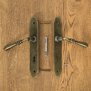 Może przedstawiać: Dwa antyczne mosiężne klamki do drzwi z otworami na klucz i ozdobnymi detalami. Klamki są zamontowane na drewnianej powierzchni i zawierają elementy montażowe.