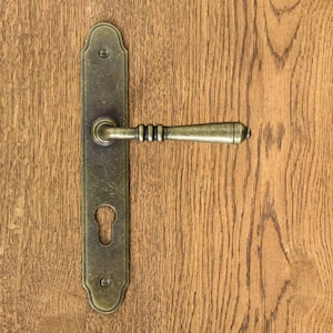 Może przedstawiać: Klamka do drzwi w stylu vintage z mosiężnym wykończeniem. Klamka ma dziurkę od klucza i ozdobną płytkę.