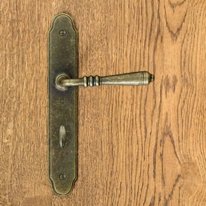 Könnte beinhalten: Ein Türgriff im Vintage-Stil mit Messing-Finish. Der Griff hat eine rechteckige Rückplatte mit einem Schlüsselloch und einen gebogenen Hebel.