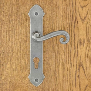 Könnte beinhalten: Ein silberfarbener Türgriff aus Metall mit einer langen, rechteckigen Grundplatte mit spitzen Enden und zwei Schraublöchern. Der Griff hat ein geschwungenes, schnörkelartiges Design, das horizontal von der Mitte der Grundplatte absteht, die zudem einen Schlüssellochausschnitt aufweist.