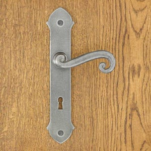 Peut inclure: Une poignée de porte en métal argenté composée d'une longue plaque rectangulaire aux extrémités pointues avec deux trous de vis circulaires. La poignée présente un design incurvé en forme de volute au-dessus d'une serrure centrale.