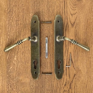 Könnte beinhalten: Ein Türgriff-Set im antiken Stil mit Messing-Finish, bestehend aus zwei langen Platten, zwei Griffen, Schrauben und einem Inbusschlüssel. Die Griffe haben ein geschwungenes Design und sind vor einem Holzgrund platziert.
