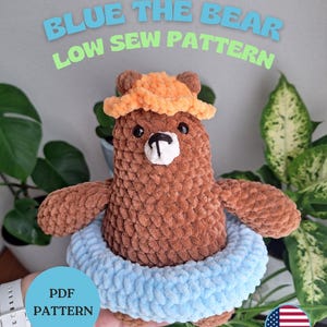 May include: A brown crocheted bear wearing an orange hat and a light blue inner tube. The text "BLUE THE BEAR LOW SEW PATTERN" is above the bear. A blue circle with "PDF PATTERN" is on the left. An American flag is on the right.