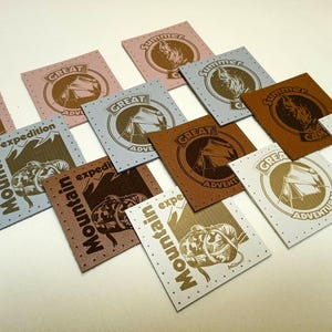 May include: A collection of square, textured labels in various colors, including pink, blue, and brown. Each label features camping-themed graphics and text such as "Great Adventures," "Summer Camp," and "Mountain Expedition."