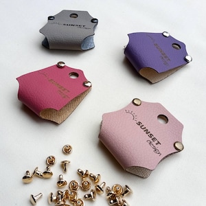 Könnte beinhalten: Vier Lederaccessoires in Grau, Lila, Rosa und Hellrosa, jeweils mit einem goldenen Niet und einem Loch. Der Text "SUNSET design" ist auf jedem aufgedruckt. Goldene Nieten sind darunter verstreut.