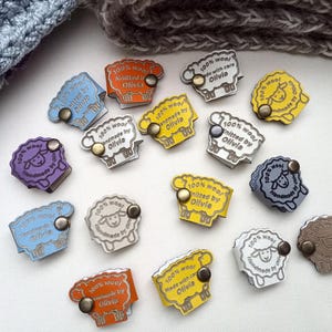 May include: A collection of colorful, sheep-shaped labels with metal fasteners. Each label features the text "100% wool" and "handmade by Olivia". The labels are in various colors, including blue, orange, yellow, and purple.