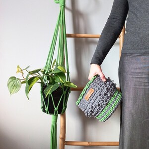 Puede incluir: Un macramé verde para colgar plantas con una planta en maceta colgando de él. Una persona sostiene una cesta de crochet gris y verde.