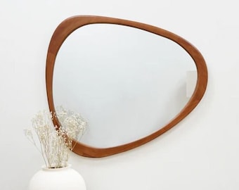 May include: A wooden, organic-shaped mirror with a light brown finish hangs on a white wall. A white vase with white flowers sits on a wooden table in front of the mirror.