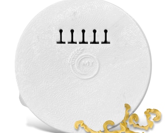 May include: White ceramic pasta bowl with a textured surface and a small circle with the letter "M" inside. The bowl has a row of six small holes on the top edge. The bowl is sitting on a white surface with a few pieces of golden pasta in the foreground.
