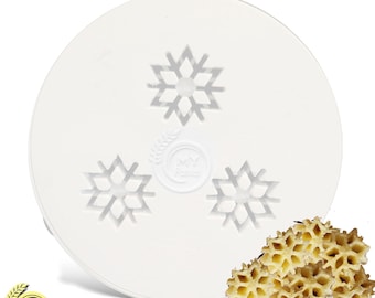 May include: White plastic pasta die with three snowflake designs. The die is used to create snowflake-shaped pasta. A pile of cooked snowflake pasta is shown in the lower right corner of the image.