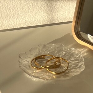 May include: Three gold bangles stacked on a clear glass dish with a textured surface.