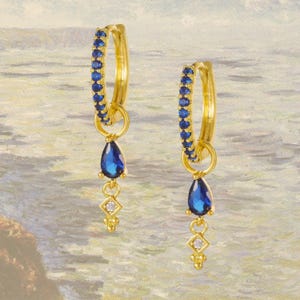 Puede incluir: Un par de pendientes de aro dorados. Cada pendiente presenta una fila de pequeñas piedras azules a lo largo del aro. Una piedra azul en forma de lágrima cuelga del aro, con un pequeño dije en forma de diamante debajo.