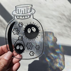 May include: A clear glass jar with a black lid and a black and white illustration of two fuzzy creatures with large eyes and small flowers inside. The jar has a cracked glass effect.