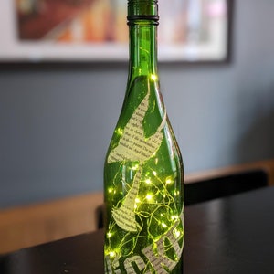 Puede incluir: Una botella de vidrio verde con un tap&oacute;n de corcho y una cadena de luces de hadas en su interior. La botella est&aacute; decorada con recortes de papel de un libro, con texto visible en la botella. La botella est&aacute; sobre una superficie oscura. #6