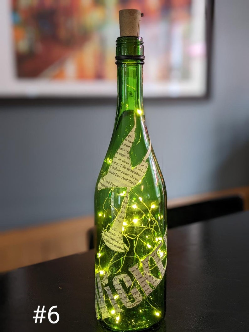 Puede incluir: Una botella de vidrio verde con un tap&oacute;n de corcho y una cadena de luces de hadas en su interior. La botella est&aacute; decorada con recortes de papel de un libro, con texto visible en la botella. La botella est&aacute; sobre una superficie oscura. #6