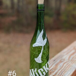 Puede incluir: Una botella de vidrio verde con un tap&oacute;n de corcho y un recorte de papel blanco de una sirena con la palabra "Nick" en la botella. La botella est&aacute; sobre una superficie de madera.