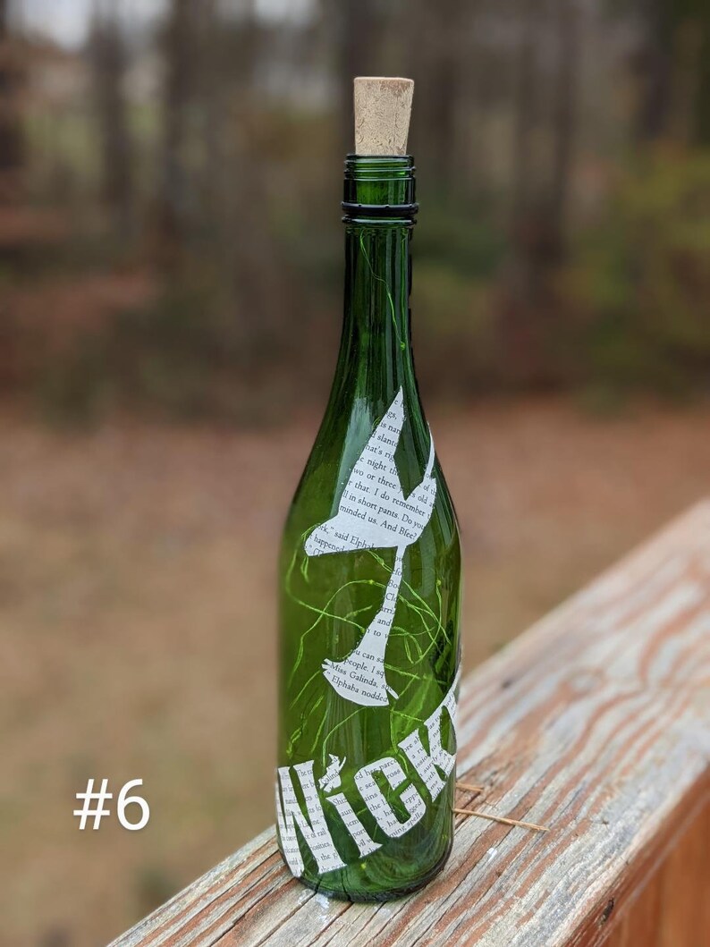 Puede incluir: Una botella de vidrio verde con un tap&oacute;n de corcho y un recorte de papel blanco de una sirena con la palabra "Nick" en la botella. La botella est&aacute; sobre una superficie de madera.