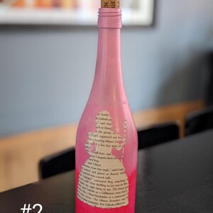 Puede incluir: Una botella de vidrio rosa con un tap&oacute;n de corcho. La botella est&aacute; decorada con una silueta de una mujer y texto de un libro. El texto dice "ay banka Galinda ach as these ha Cluach registered out lo besmetting Muses Ga few feet away hall bow, and may I enquire how you Ad Boq Mad Tibbett G from this angle" said Croppe red sofiercely that Golinder continued Bog searching h the right thing to say. The bette How thrilling to see you he Dhe Munchkinander of a commune T Galindos a Gallikinese muer fed & down Z EN LLE"