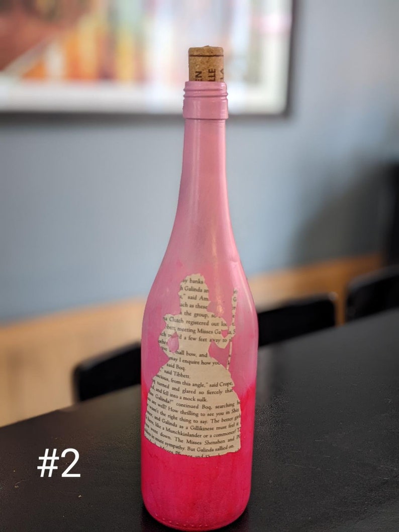 Puede incluir: Una botella de vidrio rosa con un tap&oacute;n de corcho. La botella est&aacute; decorada con una silueta de una mujer y texto de un libro. El texto dice "ay banka Galinda ach as these ha Cluach registered out lo besmetting Muses Ga few feet away hall bow, and may I enquire how you Ad Boq Mad Tibbett G from this angle" said Croppe red sofiercely that Golinder continued Bog searching h the right thing to say. The bette How thrilling to see you he Dhe Munchkinander of a commune T Galindos a Gallikinese muer fed & down Z EN LLE"