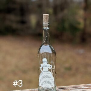 Puede incluir: Una botella de vidrio transparente con un tap&oacute;n de corcho y una silueta blanca de una mujer sosteniendo un arco y una flecha. La botella est&aacute; decorada con texto de un libro, incluyendo las palabras "Good evening, Mr. Bing." y "I am pleased to meet you, sir, more than I make it appear."
