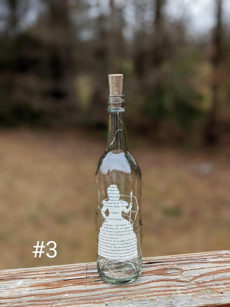 Puede incluir: Una botella de vidrio transparente con un tap&oacute;n de corcho y una silueta blanca de una mujer sosteniendo un arco y una flecha. La botella est&aacute; decorada con texto de un libro, incluyendo las palabras "Good evening, Mr. Bing." y "I am pleased to meet you, sir, more than I make it appear."