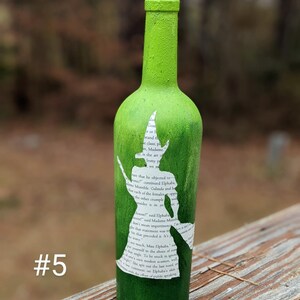 Puede incluir: Una botella de vino de vidrio verde con un tap&oacute;n de corcho. La botella est&aacute; pintada de verde y tiene una silueta blanca de una bruja con una escoba. La silueta est&aacute; hecha de una p&aacute;gina de libro con texto. La botella est&aacute; sobre una superficie de madera. #5