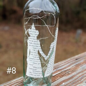 Puede incluir: Una botella de vidrio transparente con una etiqueta de papel blanco envuelta alrededor. La etiqueta tiene texto de un libro. La botella est&aacute; decorada con luces blancas de cuerda. La botella est&aacute; sentada sobre una superficie de madera.