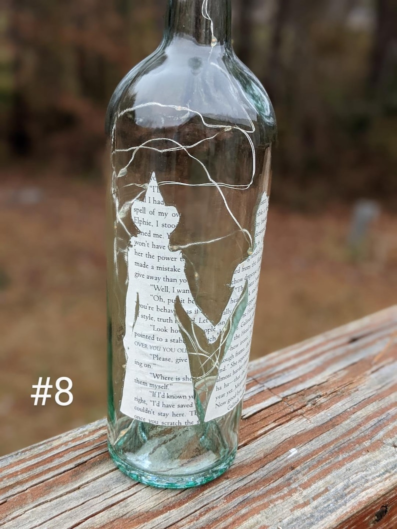 Puede incluir: Una botella de vidrio transparente con una etiqueta de papel blanco envuelta alrededor. La etiqueta tiene texto de un libro. La botella est&aacute; decorada con luces blancas de cuerda. La botella est&aacute; sentada sobre una superficie de madera.