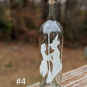 Puede incluir: Una botella de vidrio transparente con un tap&oacute;n de corcho y una silueta blanca de dos personas en su interior. La silueta est&aacute; hecha a partir de una p&aacute;gina de libro con texto visible. La botella est&aacute; sentada sobre una superficie de madera. #4