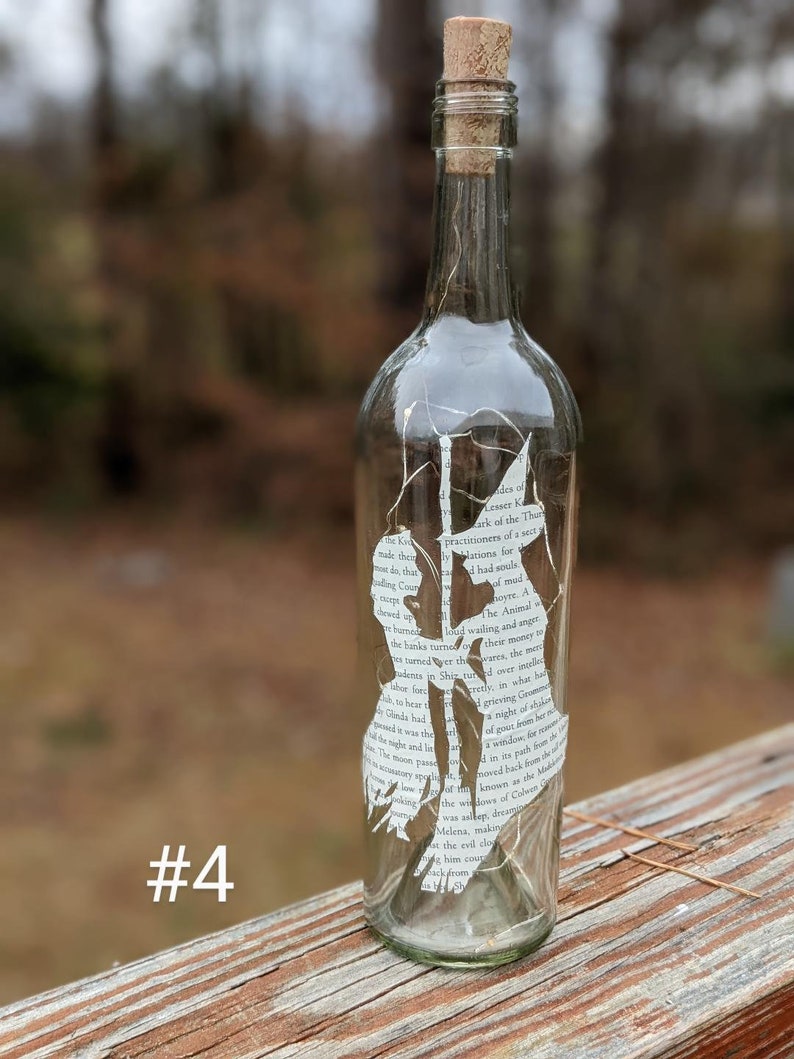 Puede incluir: Una botella de vidrio transparente con un tap&oacute;n de corcho y una silueta blanca de dos personas en su interior. La silueta est&aacute; hecha a partir de una p&aacute;gina de libro con texto visible. La botella est&aacute; sentada sobre una superficie de madera. #4