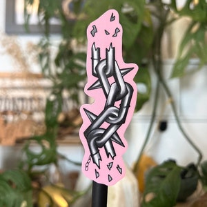 May include: A pink sticker with a broken silver chain with spikes. The chain is broken at the bottom and there are small pink pieces scattered around it.