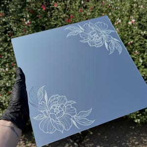 May include: A square mirror featuring white line art of floral arrangements etched into the top right and bottom left corners. The reflective surface displays a clear, smooth finish with detailed botanical illustrations of blooming flowers.