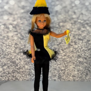 May include: A vintage doll wearing a black and yellow outfit with a matching hat. The doll has blonde hair and is holding a small yellow paper. The outfit has a black bodice and a yellow skirt with black trim. The doll is standing on a clear plastic stand.